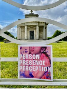 The image shows the catalogue of the portrait exhibition resting against a white iron gate with the Casino in the background above a sea of green grass. The catalogue has the image of a child on the front cover, in hues of pink, with the name of the exhibition across the face in white. It is called Person, Presence, Perception